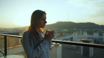 mooi vrouw drankjes kop van koffie of thee, aan het kijken een mooi stedelijk visie en genieten kom tot rust ademen vers bevriezing lucht Aan balkon Bij dageraad video