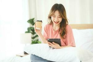 Asian woman with coffee using digital tablet for social message telemedicine and mental health or shopping online on at bedroom morning time photo