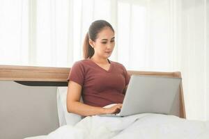 Young adult asian woman using laptop on bed for telemedicine mental health or shopping online photo