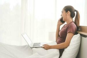 Asian woman using laptop and talk with smartphone on bed for telemedicine mental health at home photo