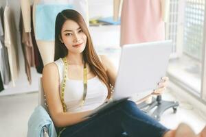 Beautiful young adult asian woman tailor designer sitting using laptop and eye looking camera in workshop photo