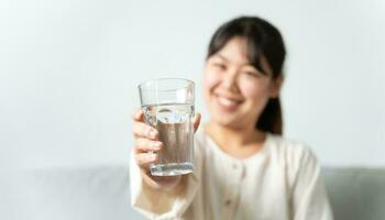 mujer joven sana y hermosa sosteniendo un vaso de agua sentado en el sofá en la sala de estar. foto