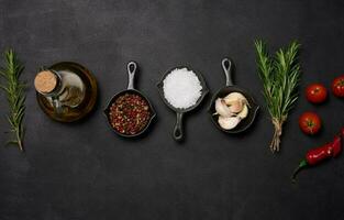 miniatura sartenes con especias, sal, negro pimienta y fragante pimienta, un puntilla de Romero en un negro mesa. especias para Cocinando foto