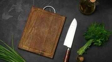 Empty wooden board, green onions, bunch of dill and parsley on a black table, top view photo