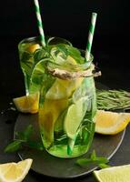 Lemonade in a transparent glass with lemon, lime, rosemary sprigs and mint leaves on a black background photo