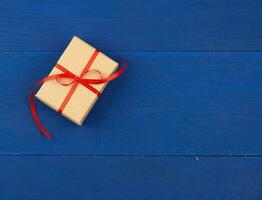 gift wrapped in brown kraft paper and tied with a thin red ribbon on a blue wooden background, top view, flat lay. Eco package design photo