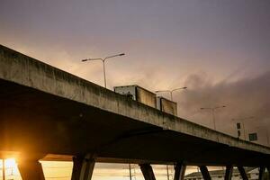 Bottom view of elevated concrete highway with truck driving. Overpass concrete road. Road flyover structure. Motorway with sunset sky. Concrete bridge engineering construction. Bridge architecture. photo