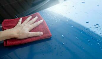 Car wash service. Man hand holding red microfiber cloth polish blue car and water drop after cleaning. Auto care service business concept. Man cleaning and detailing luxury car with microfiber cloth. photo