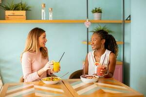 Young black and caucasian woman having good time, drinking fresh juices and having healthy breakfast in the cafe photo