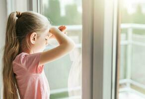 triste pequeño niña mirando mediante ventana a hogar. foto