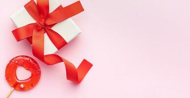 Top view photo of gift box, heart shaped lollipop on pastel pink background with copy space in the middle.