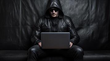 Portrait of Causation Young Man Wearing Goggles with Black Leather Jacket and Working on Laptop on Sofa. . photo