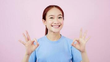 mignonne asiatique femme dans bleu T-shirt à la recherche à caméra et donnant D'accord signe sur rose Contexte video