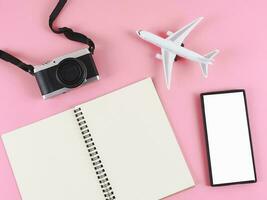 flat lay of blank paper note book, blank white screen mobile phone,  airplane model  and camera on pink background. Travel plan concept. photo