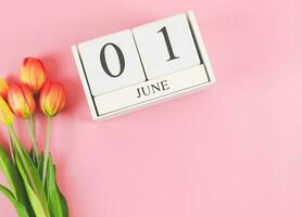 flat lay of wooden calendar with date June 01 on pink  background with orange and yellow tulips, copy space. photo