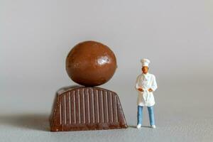 miniatura personas cocinero con chocolate en pie mientras en pie en contra un gris antecedentes foto