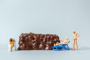 Miniature people wearing swimsuit relaxing on chocolate bars on gray background. World Chocolate Day Concept photo