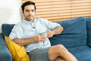 Rest Concept. Happy guy drinking coffee sitting on comfortable couch at home in living room. a smiling young bearded man drinking tea from a mug at the living room. photo