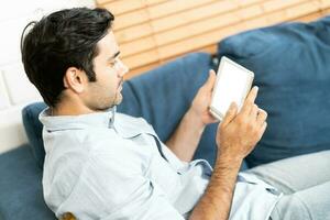 Handsome man is using a digital tablet while resting on couch at home. man sitting on sofa and holding tablet computer with isolated screen photo