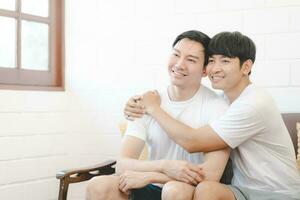 Happy Asian gay couple hug together on sofa. Asian LGBT couple embracing together at home. Diversity of LGBT relationships. A gay couple concept. LGBT multi relationship. photo