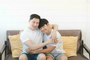 Happy Asian gay couple hug together on sofa. Asian LGBT couple embracing together at home. Diversity of LGBT relationships. A gay couple concept. LGBT multi relationship. photo