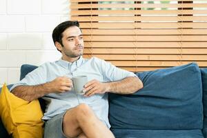 Rest Concept. Happy guy drinking coffee sitting on comfortable couch at home in living room. a smiling young bearded man drinking tea from a mug at the living room. photo