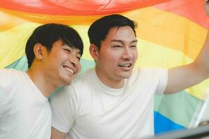 Happy Asian LGBT couple, sitting on the sofa holding and waving rainbow LGBT Pride flag together in the bedroom at home. Diversity of LGBT relationships. A gay couple concept. Loving gay couple. photo