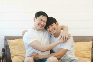 Happy Asian gay couple hug together on sofa. Asian LGBT couple embracing together at home. Diversity of LGBT relationships. A gay couple concept. LGBT multi relationship. photo