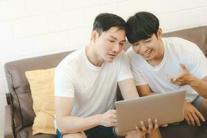 asiático gay Pareja utilizando computadora para vídeo conferencia en sofá. alegre joven gay Pareja sonriente alegremente mientras compras en línea a hogar. foto