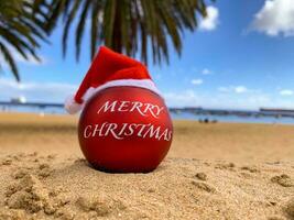 bomba de navidad en el sombrero de santa en la playa tirada en la arena con palmeras y cielo azul en el fondo. feliz navidad desde el paraíso, isla exótica. hawai, islas canarias, bali. foto