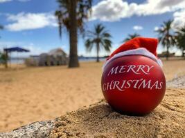 Christmas bomb in Santa's hat on the beach lying on the sand with palm trees and blue sky on the background. Merry christmas from paradise, exotic island. Hawaii, Canary islands, Bali. photo