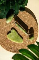 Jade Gua sha scraper and face roller massager on a cork round stand with a monstera leaf. photo