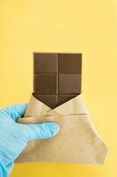 bar de Leche chocolate en un envoltura es retenida por un mano en un médico guante en un amarillo antecedentes. dulzura. mundo chocolate día 2020 durante el coronavirus. foto