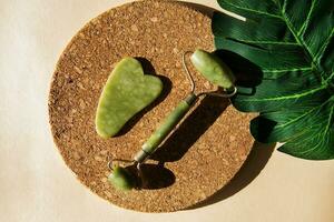 Jade Gua sha scraper and face roller massager on a cork round stand with a monstera leaf. photo