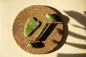 Jade Gua sha scraper and face roller massager on a cork round stand with a monstera leaf. photo