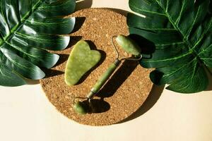 Jade Gua sha scraper and face roller massager on a cork round stand with a monstera leaf. photo