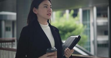 metraggio di contento giovane asiatico donna d'affari nel completo da uomo Tenere un' il computer portatile e tazza mentre a piedi nel un' moderno attività commerciale costruzione. attività commerciale e persone concetti. video