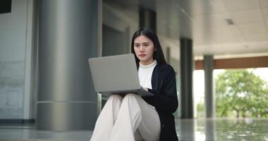 Aufnahmen von jung elegant asiatisch beschäftigt Geschäft Frau im ein passen Arbeiten mit ein Laptop während Sitzung auf Treppe im Vorderseite von ein modern Geschäft Gebäude. Geschäft und Menschen Konzepte. video