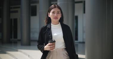 Footage of Young smiling elegant Asian busy business woman in a suit holding a cup of coffee and walking in front of a modern business building. Business and people concepts. video