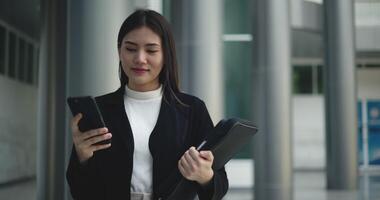 voorkant visie schot, beeldmateriaal van jong glimlachen elegant Aziatisch bezig bedrijf vrouw in een pak gebruik makend van een smartphone en wandelen in voorkant van een modern bedrijf gebouw. bedrijf en mensen concepten. video