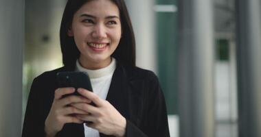 Front view shot, Footage of Young smiling elegant Asian busy business woman in a suit using a smartphone and walking in front of a modern business building. Business and people concepts. video