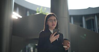 imágenes de joven asiático ocupado mujer de negocios vistiendo lentes en un traje participación un taza de café y hablando en el teléfono mientras en pie en un moderno negocio edificio. negocio y personas conceptos. video