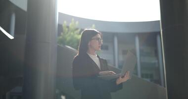 Footage of Young elegant Asian busy businesswoman wearing glasses in a suit using a laptop while standing in front of a modern business building. Business and people concepts. video