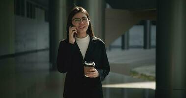 imágenes de joven asiático ocupado mujer de negocios vistiendo lentes en un traje participación un taza de café y hablando en el teléfono mientras caminando en frente de un moderno negocio edificio. negocio y personas conceptos. video