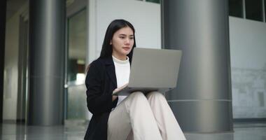 beeldmateriaal van jong elegant Aziatisch bezig bedrijf vrouw in een pak werken met een laptop terwijl zittend Aan trap in voorkant van een modern bedrijf gebouw. bedrijf en mensen concepten. video