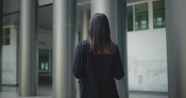 retour vue tir, métrage de Jeune souriant élégant asiatique occupé affaires femme dans une costume en utilisant une téléphone intelligent et en marchant dans de face de une moderne affaires bâtiment. affaires et gens concepts. video