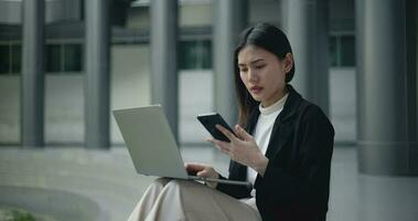 Aufnahmen von jung elegant asiatisch beschäftigt Geschäft Frau im ein passen Arbeiten mit ein Laptop und reden auf Handy, Mobiltelefon während Sitzung auf Treppe im Vorderseite von ein modern Geschäft Gebäude. Geschäft und Menschen Konzepte. video