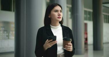Footage of Young elegant Asian busy business woman in a suit holding mobile phone and a cup of coffee while standing in front of a modern business building. Business and people concepts. video