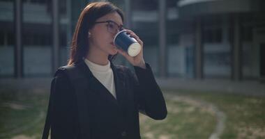 beeldmateriaal van jong glimlachen elegant Aziatisch bezig zakenvrouw vervelend bril in een pak drinken koffie terwijl staand in voorkant van een modern bedrijf gebouw. bedrijf en mensen concepten. video