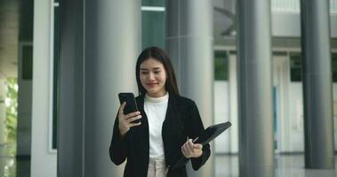 de face vue tir, métrage de Jeune souriant élégant asiatique occupé affaires femme dans une costume en utilisant une téléphone intelligent et en marchant dans de face de une moderne affaires bâtiment. affaires et gens concepts. video